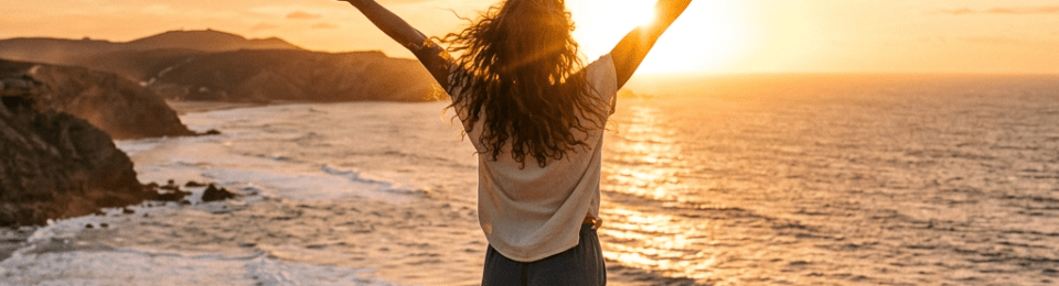 Woman standing barefoot on cliff with arms raised facing ocean sunset